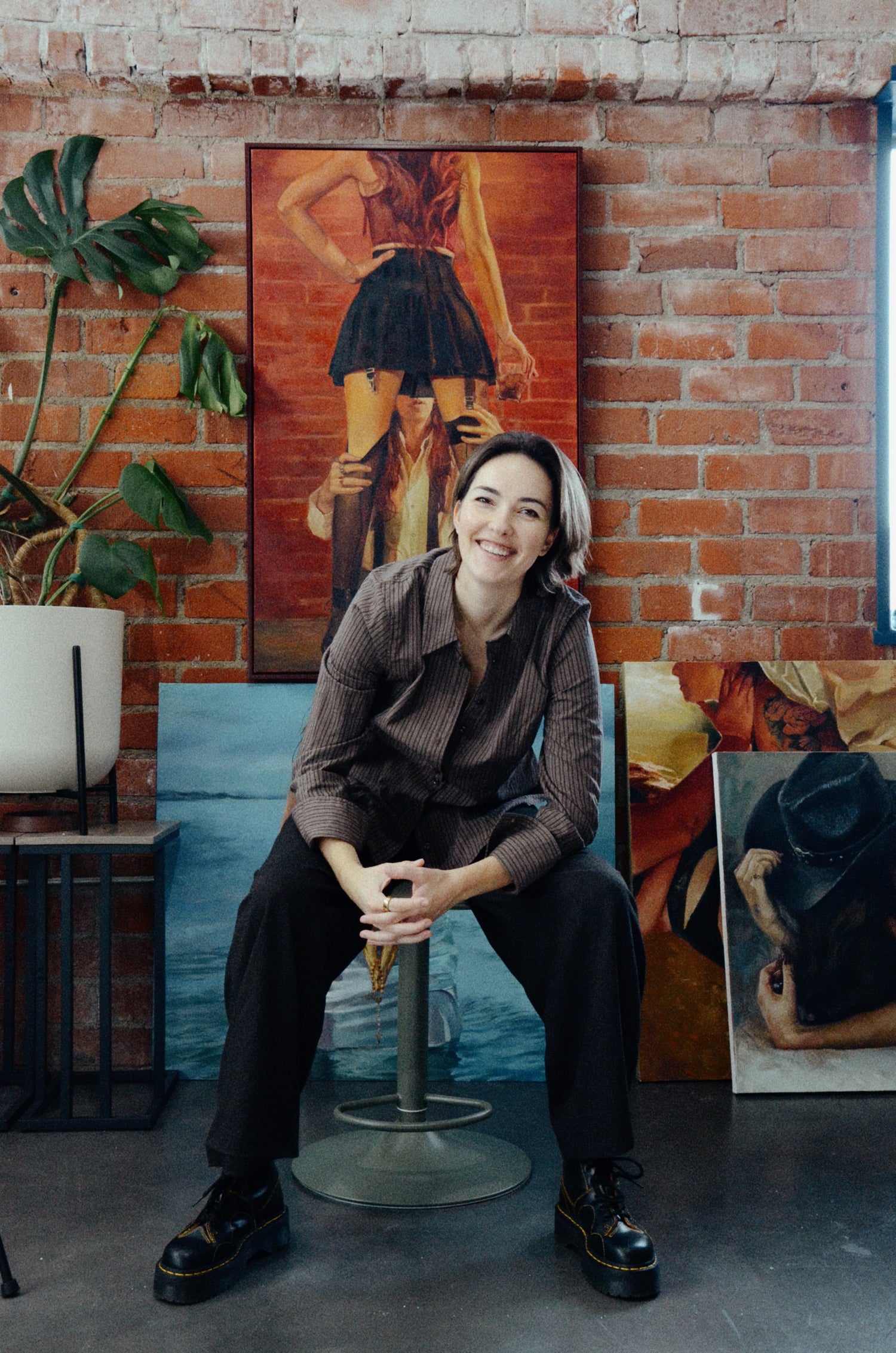Woman sitting on a stool in front of artistic wall decorations in an indoor setting.