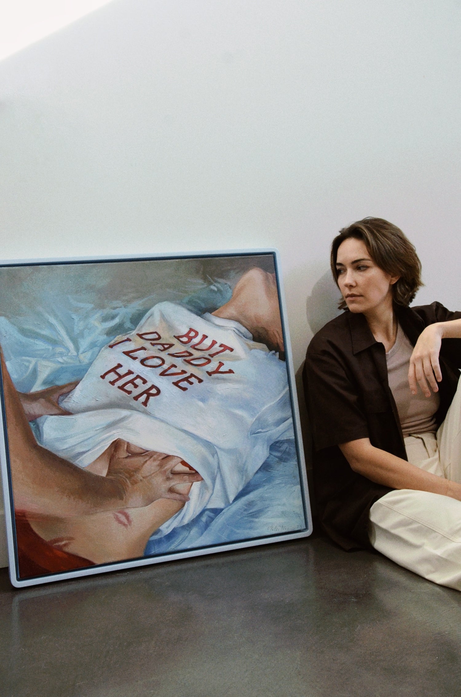 Halie Torris sitting next to her "but daddy i love her" oil painting with cropped hair.
